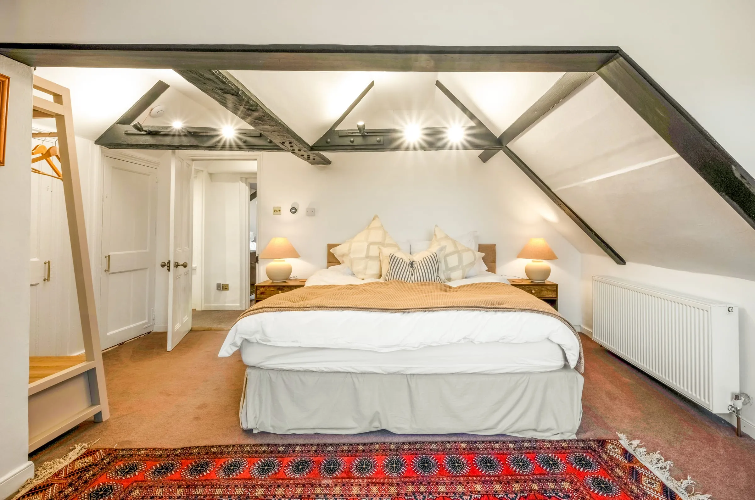 Bedroom under exposed beams with a king-size bed, white linen and a red patterned rug