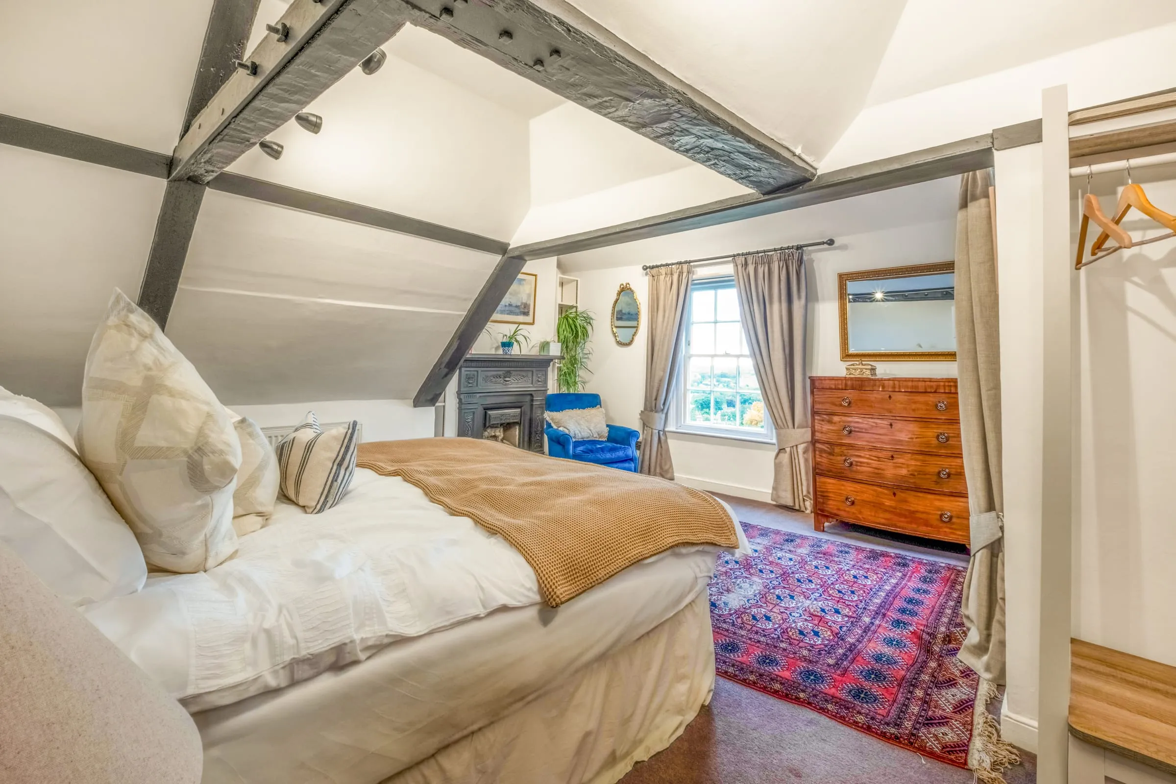Bedroom with beamed ceiling, chest of drawers, fireplace and a window looking towards the trees