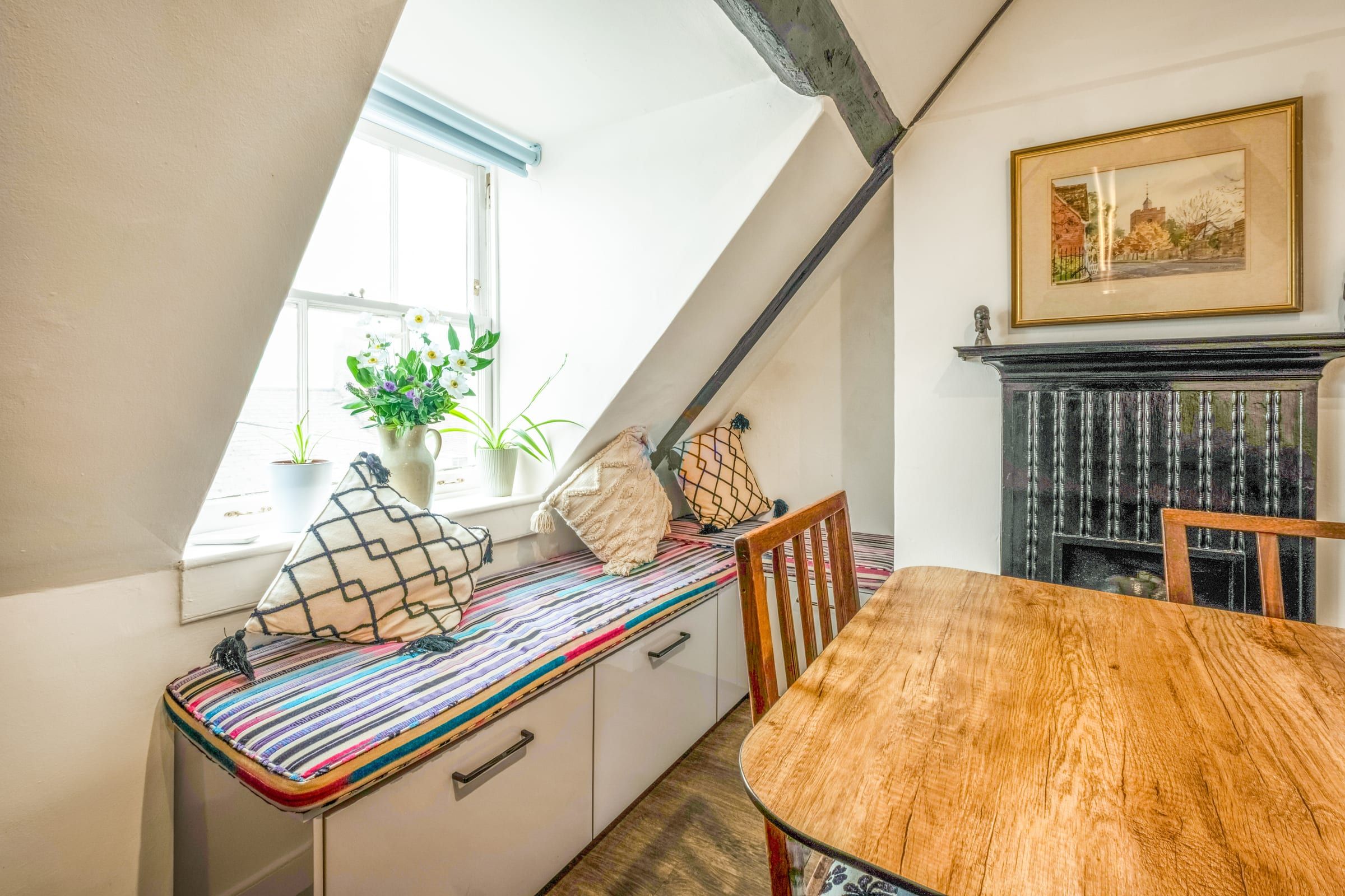 Wooden dining table beside a cushioned window seat with a black cast-iron fireplace in the corner